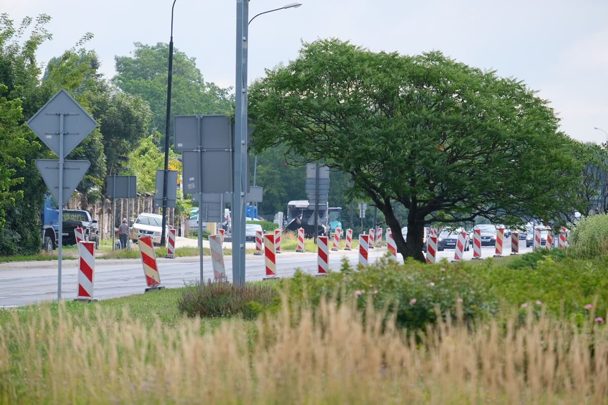 Pachołki na pasie zieleni wzdłuż al. Tysiąclecia, a w oddali maszyna do układania asfaltu